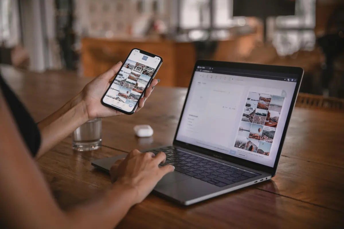 A person using a laptop and holding a smartphone, both screens displaying a grid of photos.