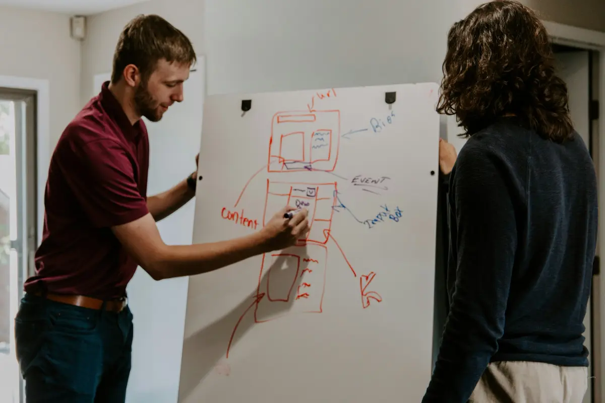 A man draws a website wireframe on a whiteboard while another person observes.